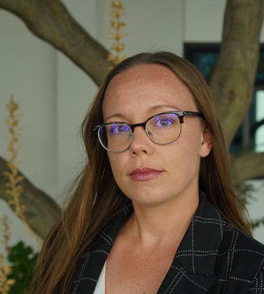 Woman with glasses and dark plaid blazer, neutral expression, standing by tree and windows