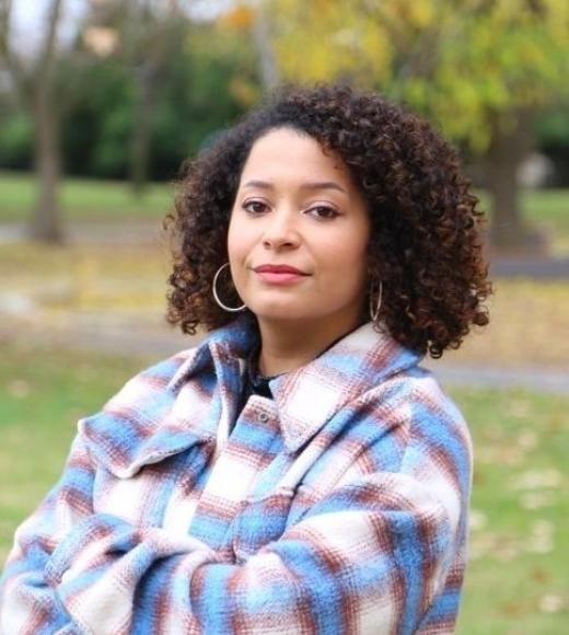 A woman with curly hair stands outdoors in a park setting with trees and autumn foliage, wearing a plaid jacket, looking at the camera.