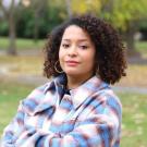 A woman with curly hair stands outdoors in a park setting with trees and autumn foliage, wearing a plaid jacket, looking at the camera.