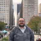 Manny standing in downtown Chicago, framed by autumn trees and towering city buildings. Wearing a gray camo hoodie and glasses, he’s captured in a lively urban plaza with people strolling and a blue kiosk in the background.