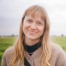 Smiling young woman with long blond hair and bangs in beige fleece, grassy field background